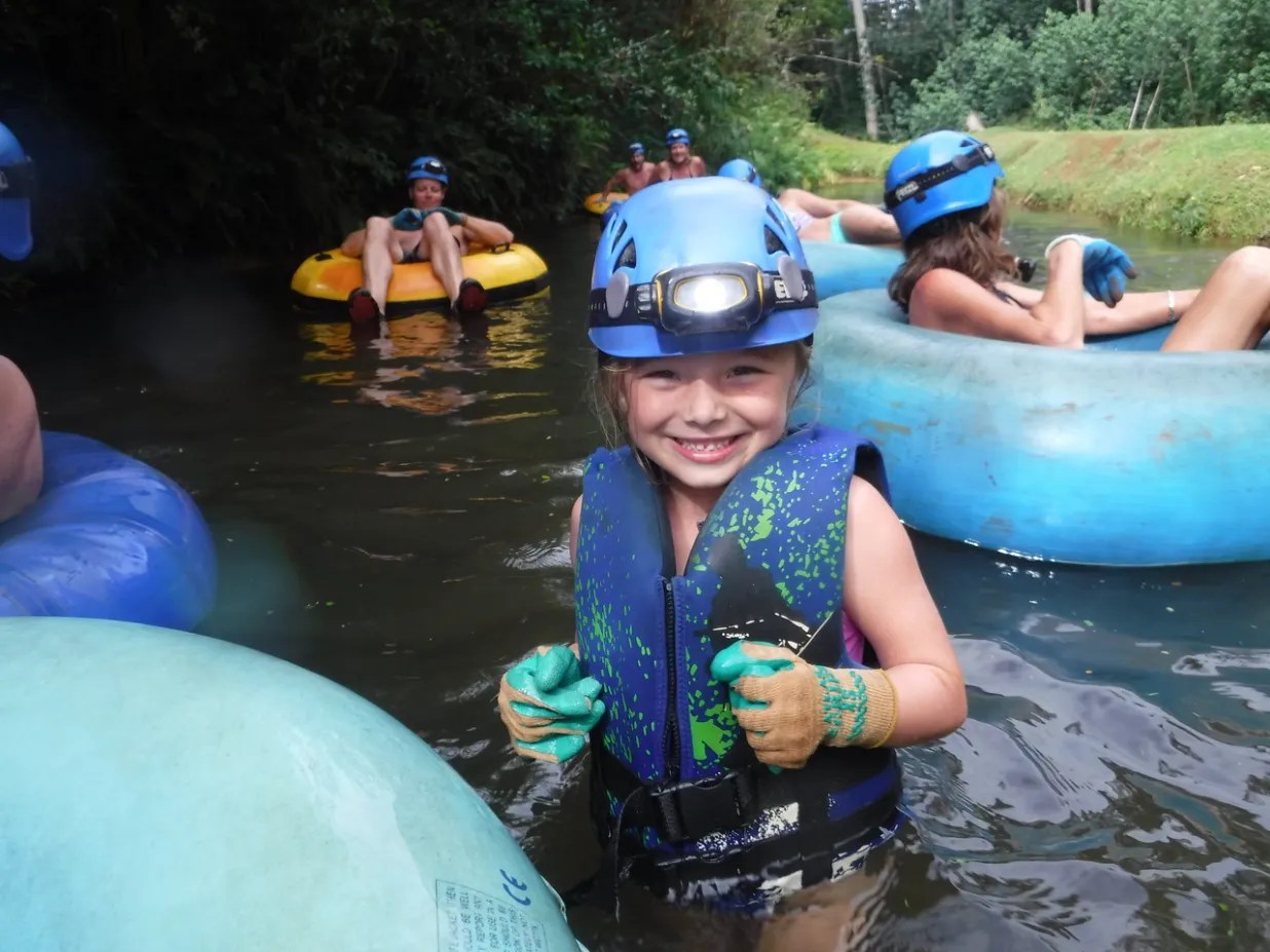 Young girl in a life jacket and helmet smiling while tubing in a river with others.