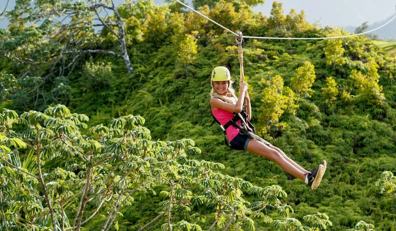 Person ziplining through lush green forest, wearing helmet and safety gear.