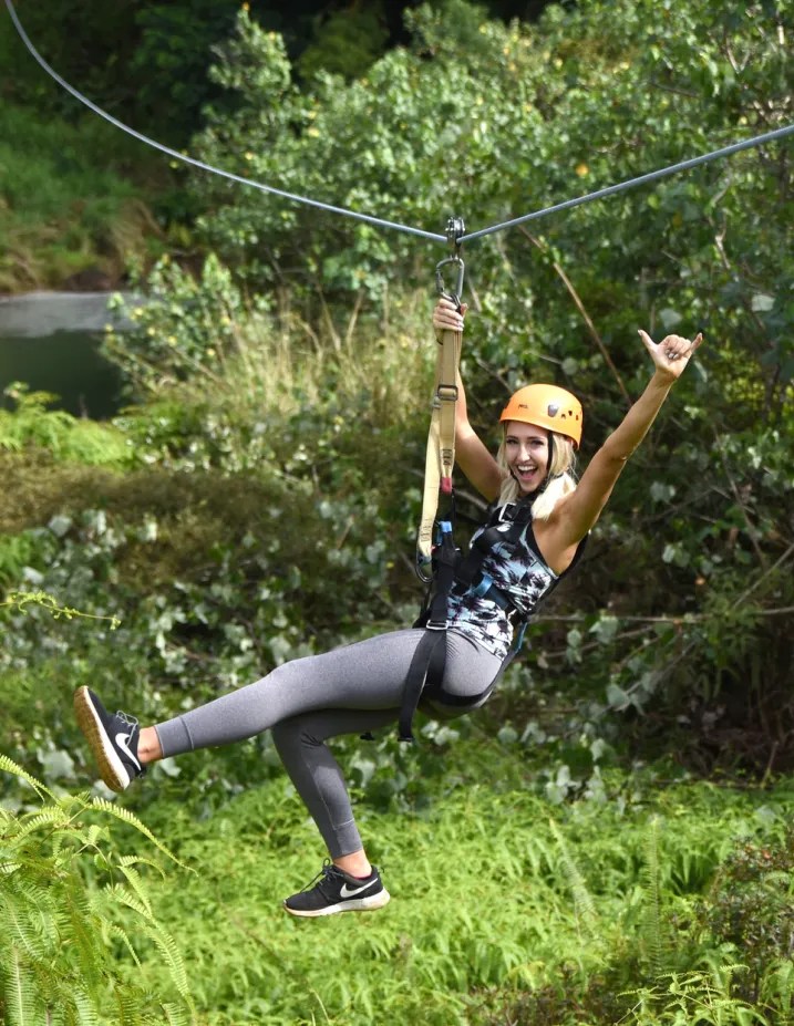 Person ziplining through a green forest area, smiling and giving a thumbs-up.