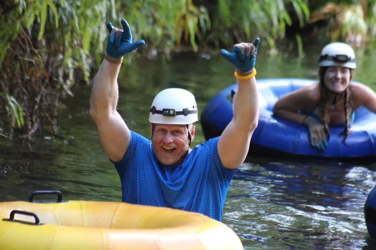 Two people tubing in a river, wearing helmets and gloves, surrounded by greenery.