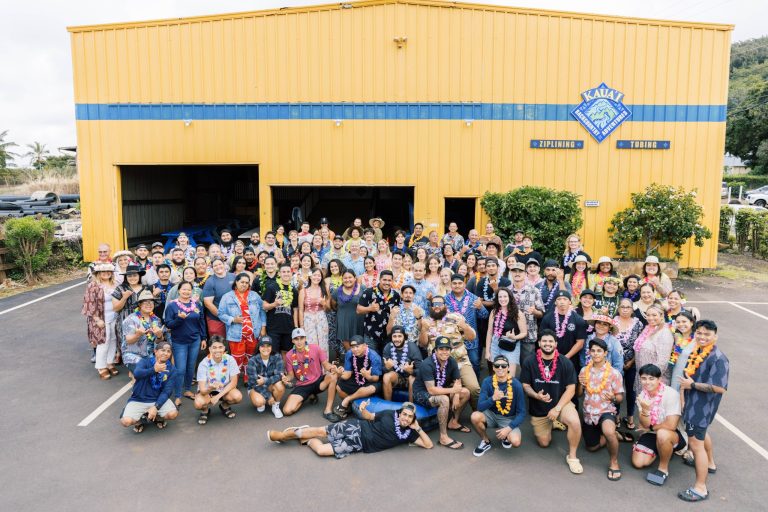 Large group in colorful attire poses outside a yellow building.