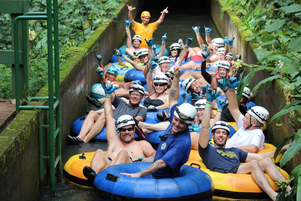 Group of people in helmets tubing down a canal, cheering and posing for a photo.