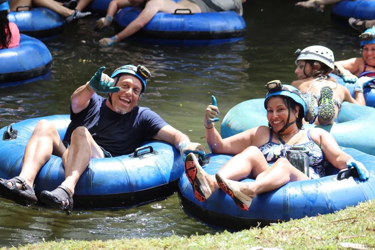 Two people in helmets and gloves tubing on a river, smiling and giving thumbs up.