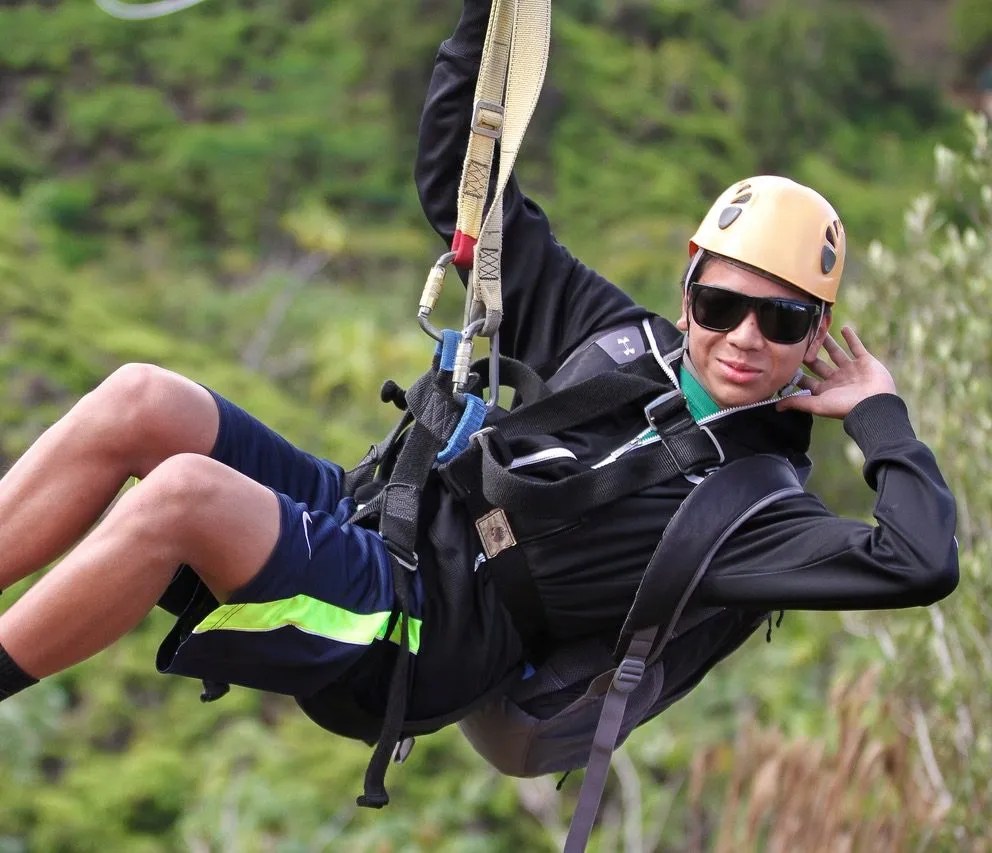 Person zip-lining, wearing helmet and sunglasses, in outdoor setting.