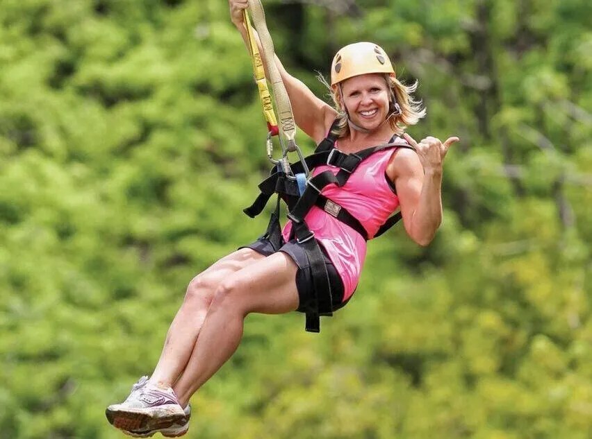 Woman wearing a helmet and harness smiling while zip-lining through a forested area.