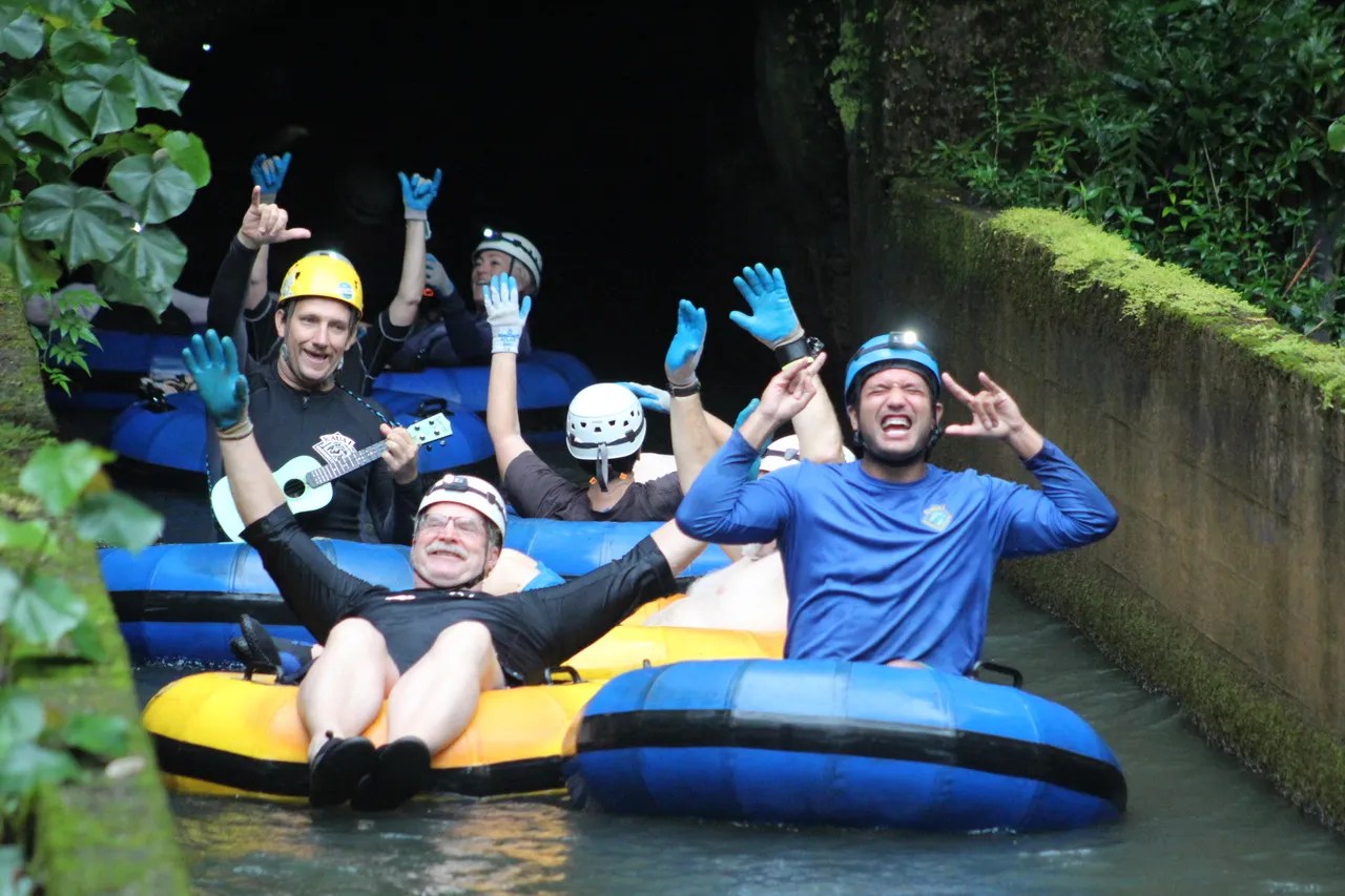 People wearing helmets and gloves tube down a water passage, some playing instruments and making gestures.