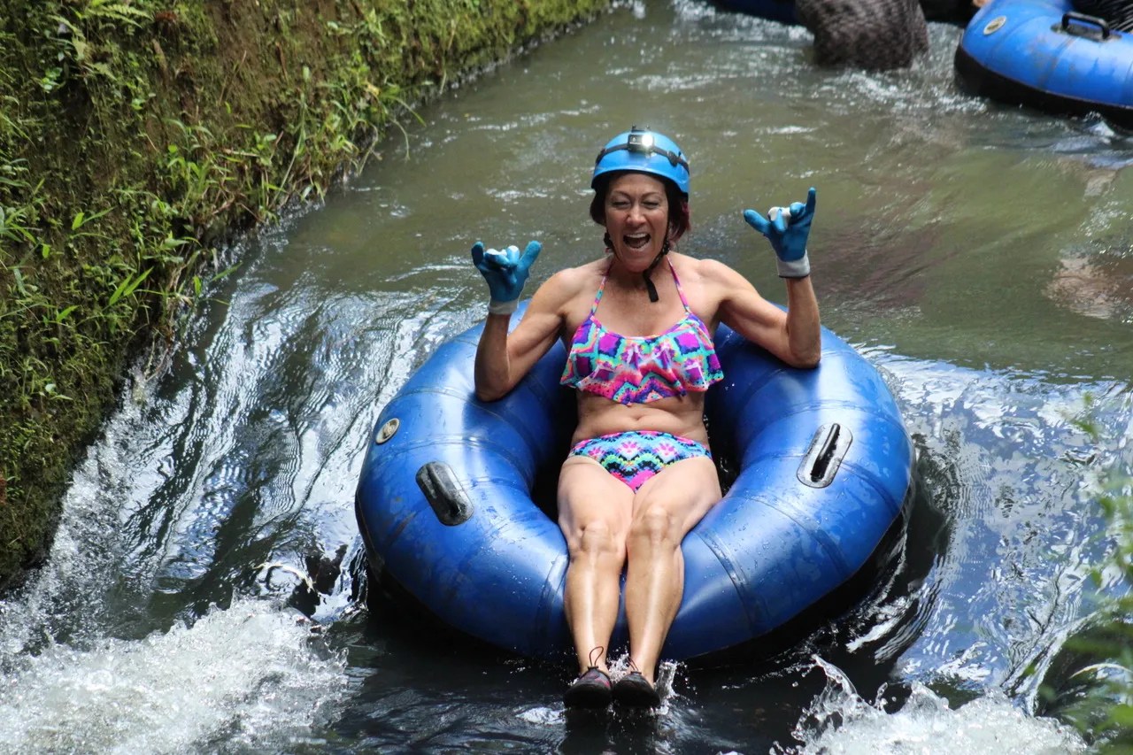 Person tubing on a narrow river, wearing a helmet and colorful swimsuit, raising both hands with excitement.