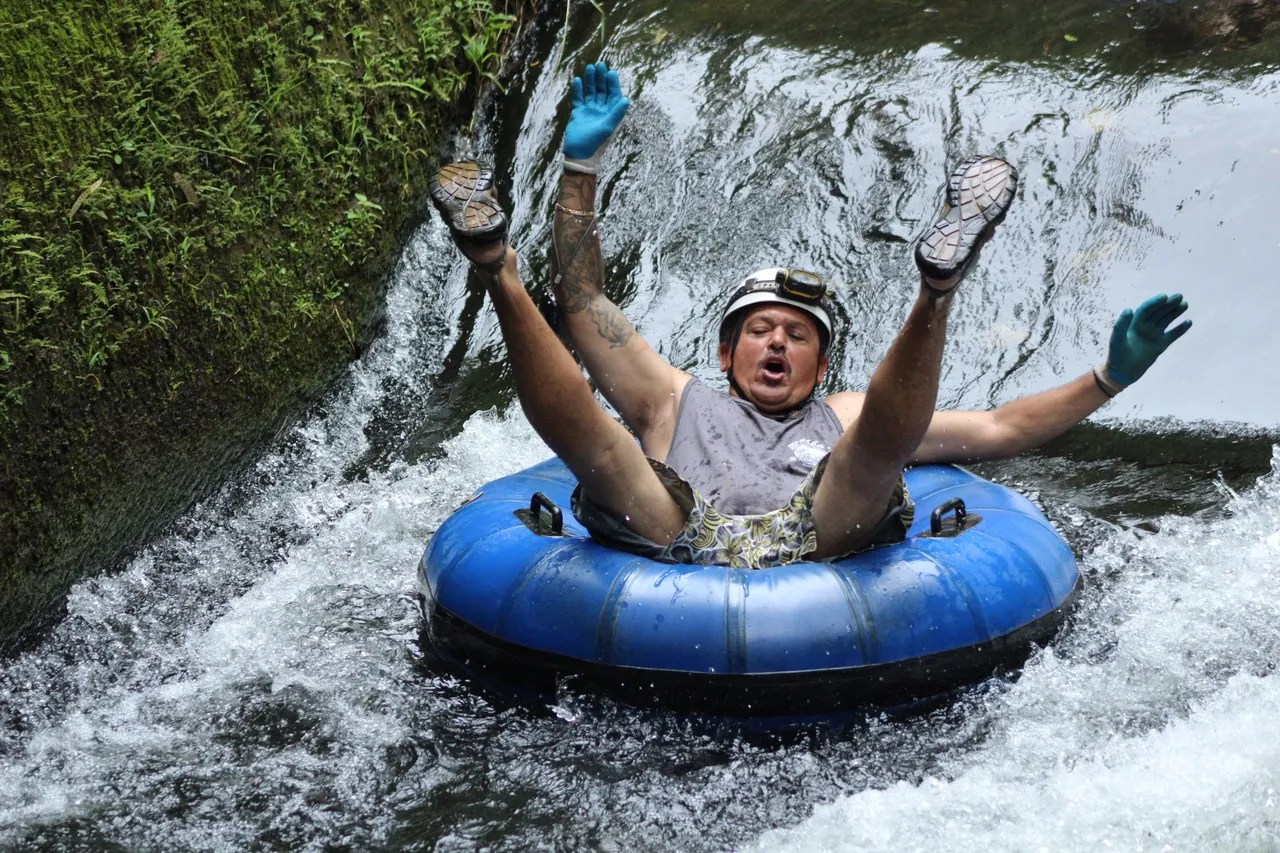 Person tubing down a river, wearing gloves and a helmet, with arms and legs raised.