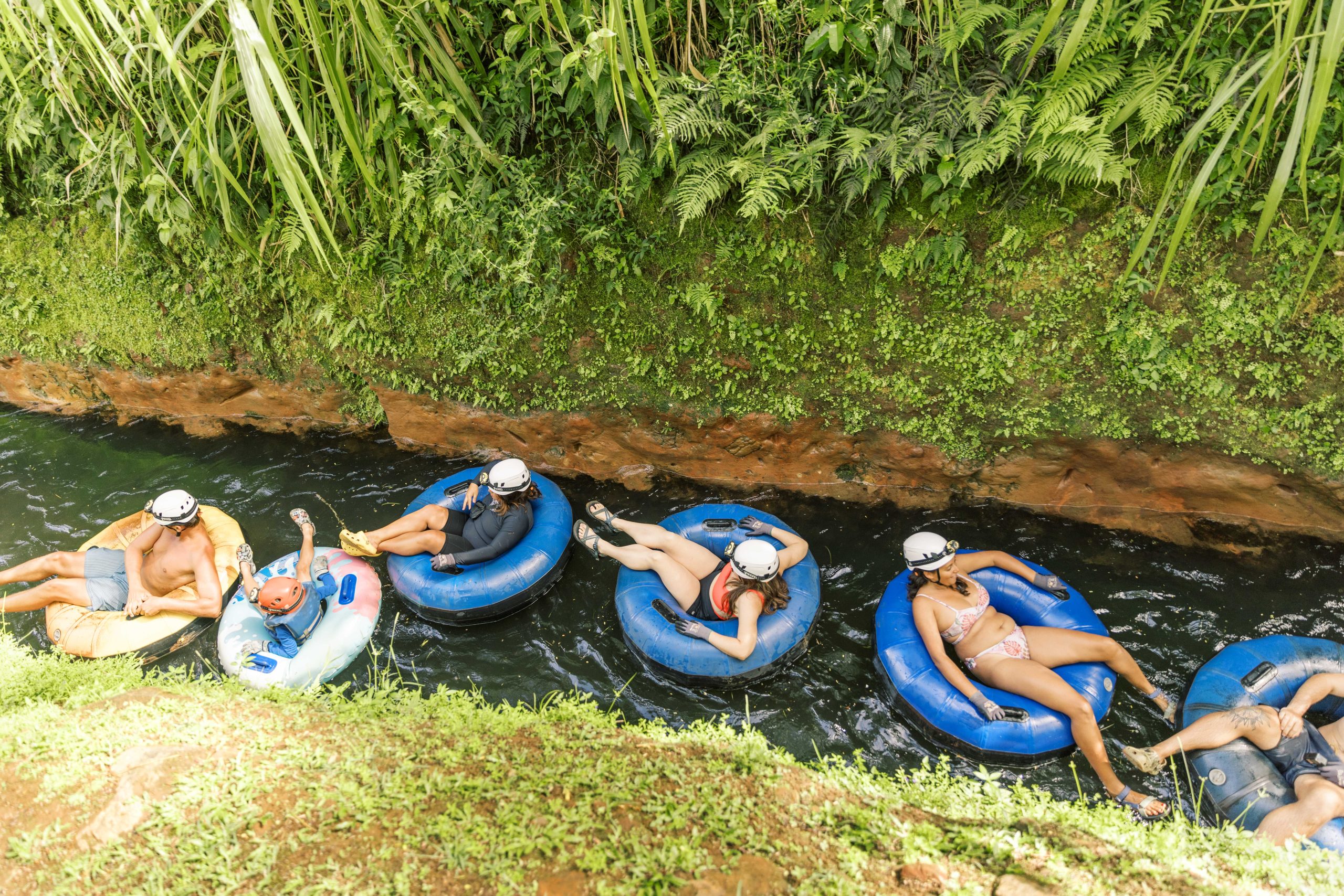 People in helmets relaxing on inflatable tubes in a river surrounded by greenery.