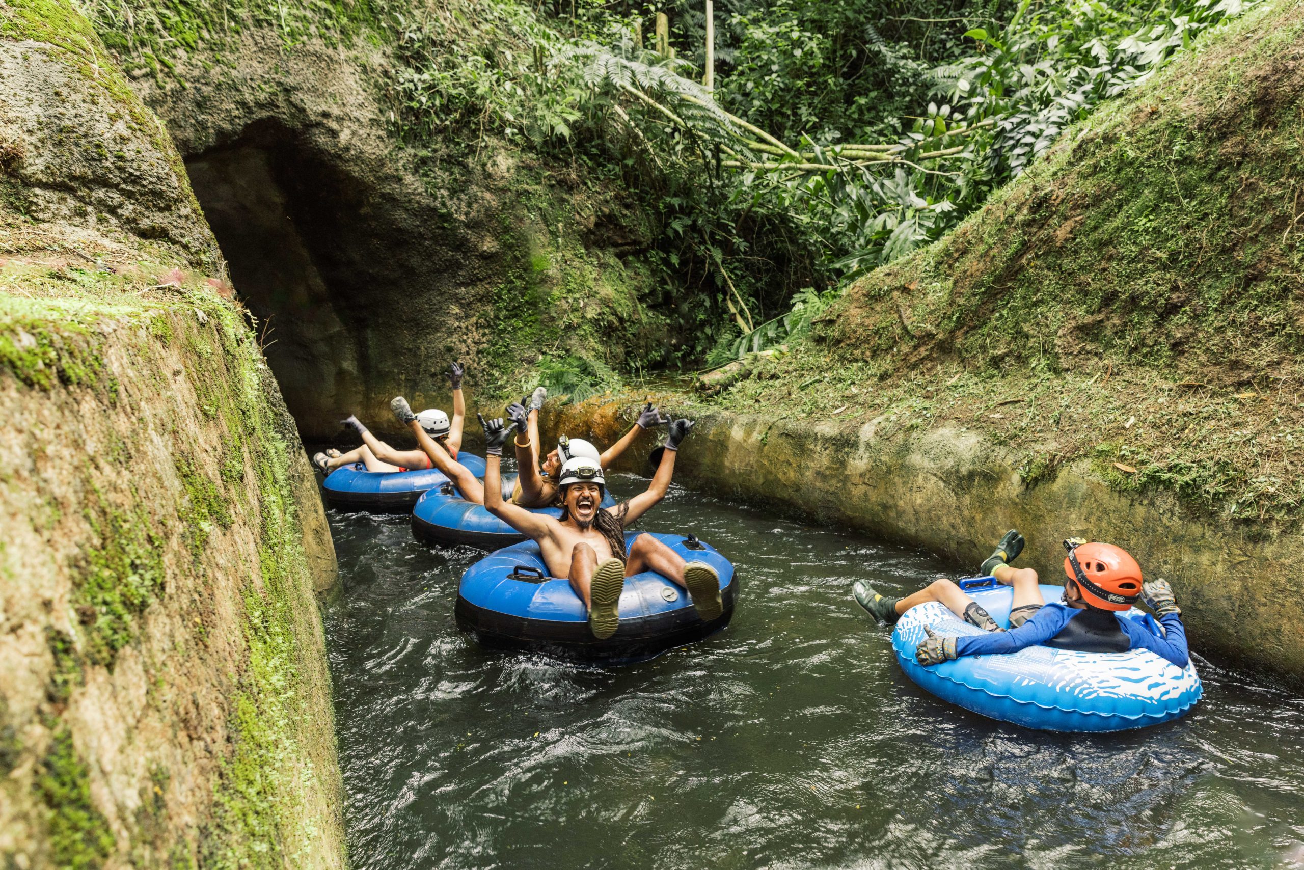People tubing with helmets in a narrow jungle waterway.