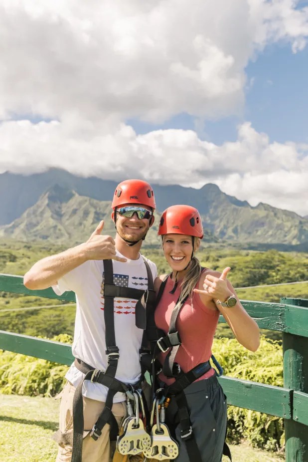 Two people in helmets giving thumbs up with mountain scenery.