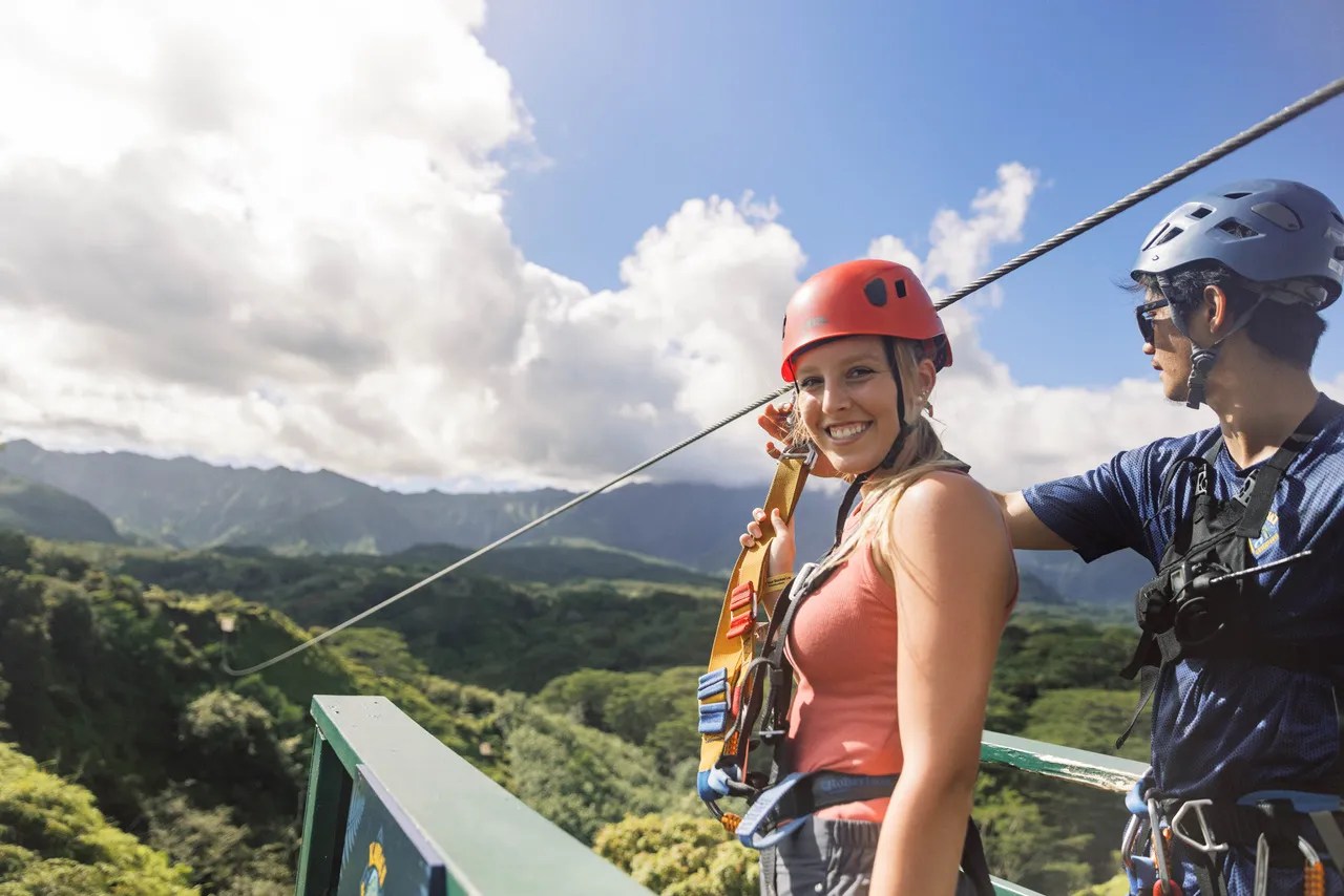Two people in helmets ready to zip line over lush green landscape.