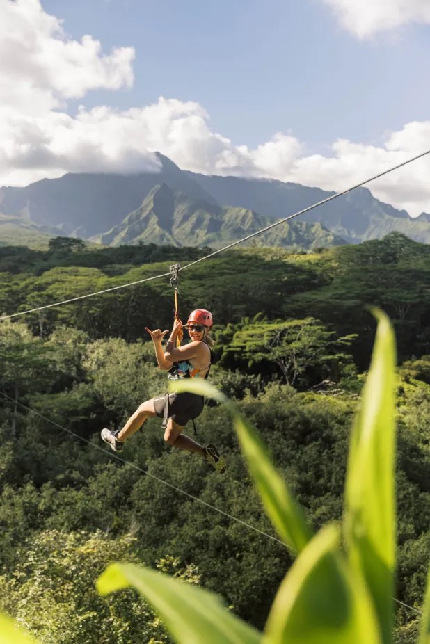 Person zip-lining over lush green forest with mountains in the background.