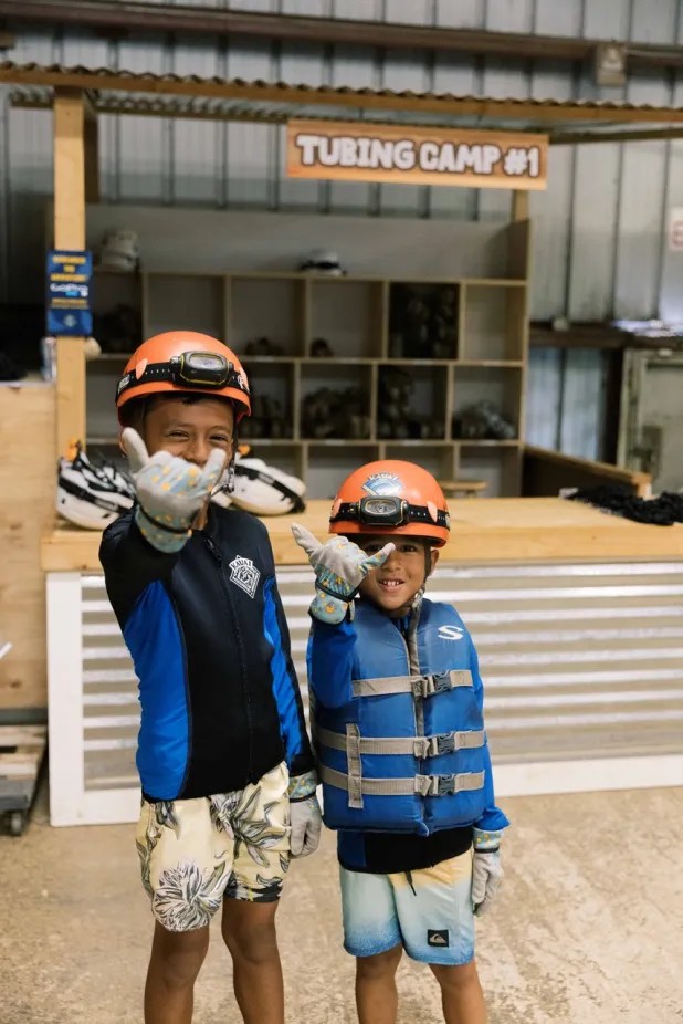 Two kids in helmets and gear smiling and gesturing at a tubing camp.