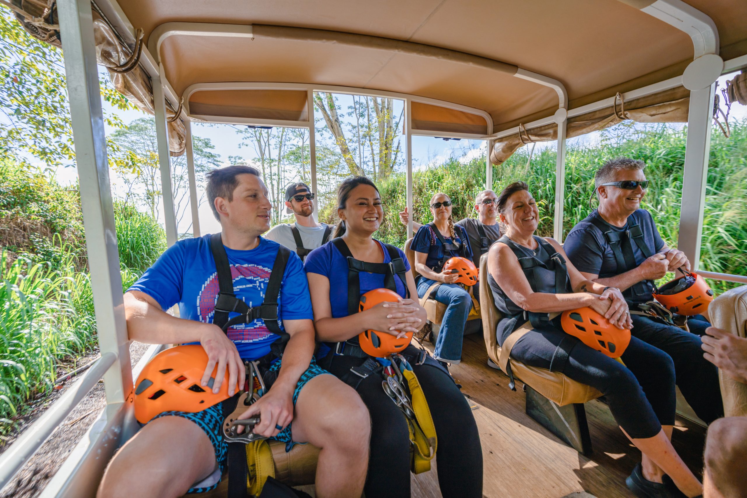Group of people seated in an open-air vehicle, holding orange helmets, surrounded by greenery.