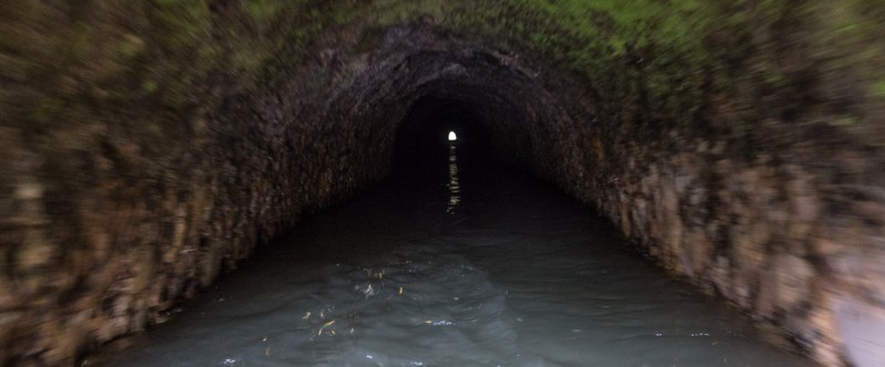 Dark, narrow tunnel with water flowing through it; light visible at the end.