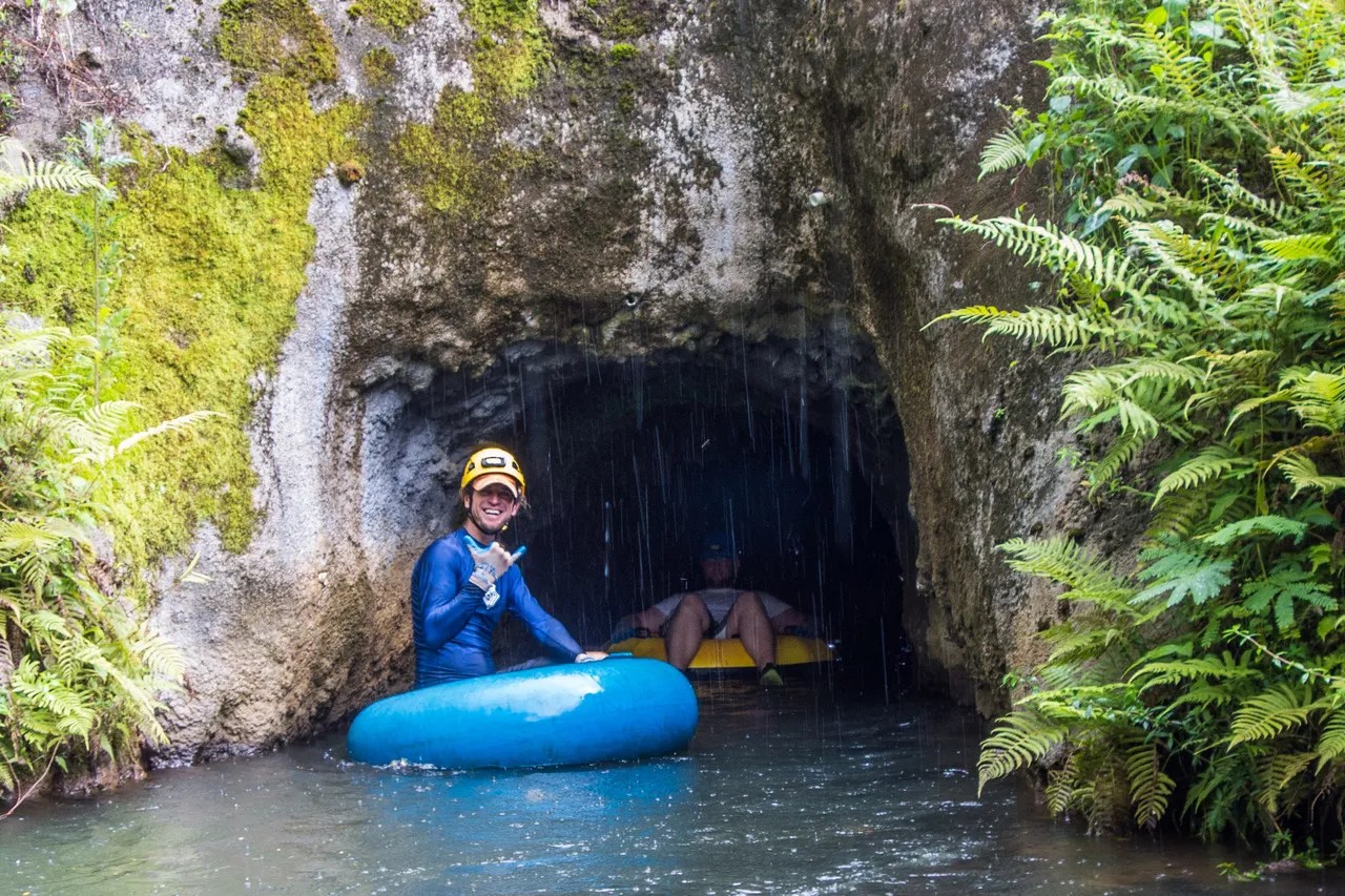 Two people tubing out of a cave, surrounded by moss and ferns.