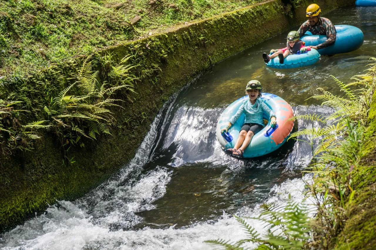 People tubing down a narrow, lush green water canal.