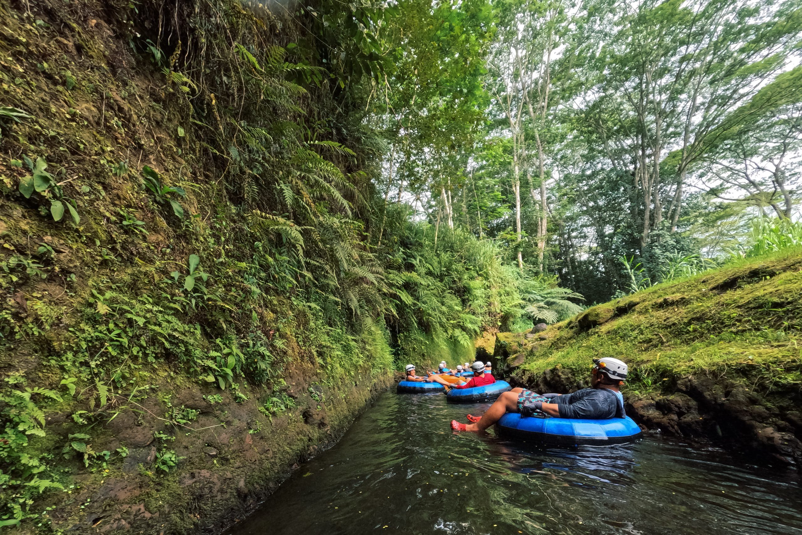 People river tubing through a lush, narrow stream surrounded by greenery.