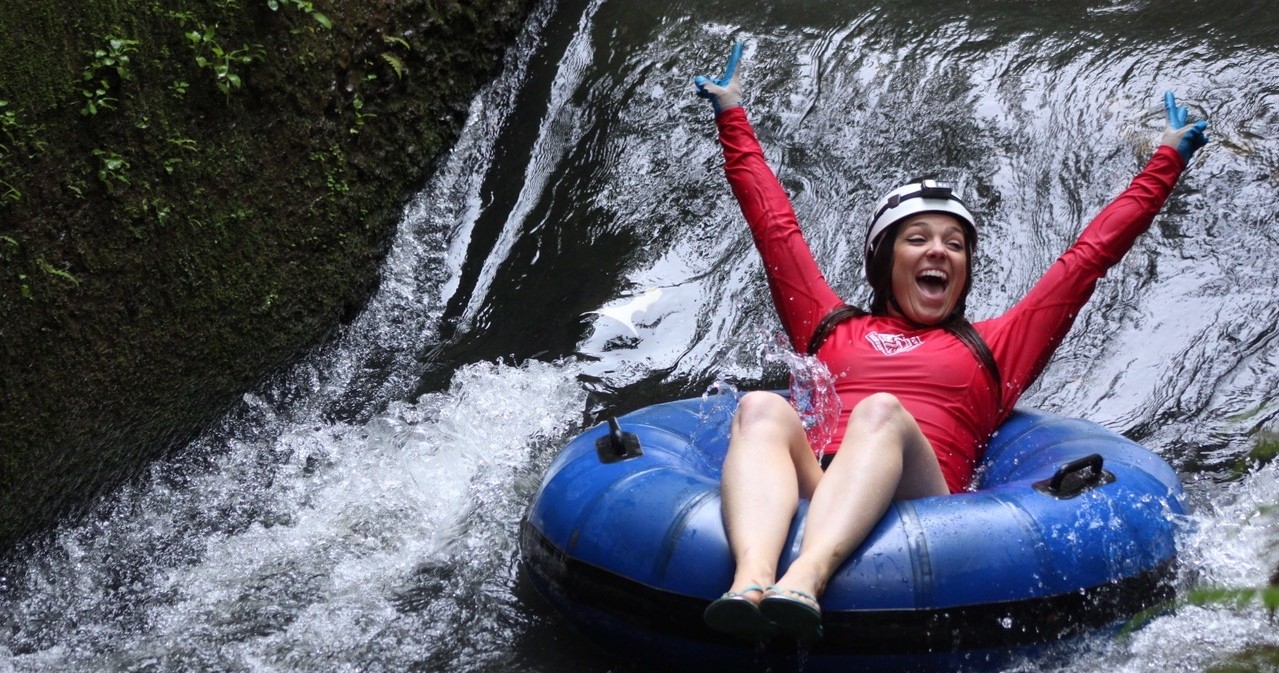 Person riding an inflatable tube down a water slide, smiling with arms raised, wearing a helmet and red top.