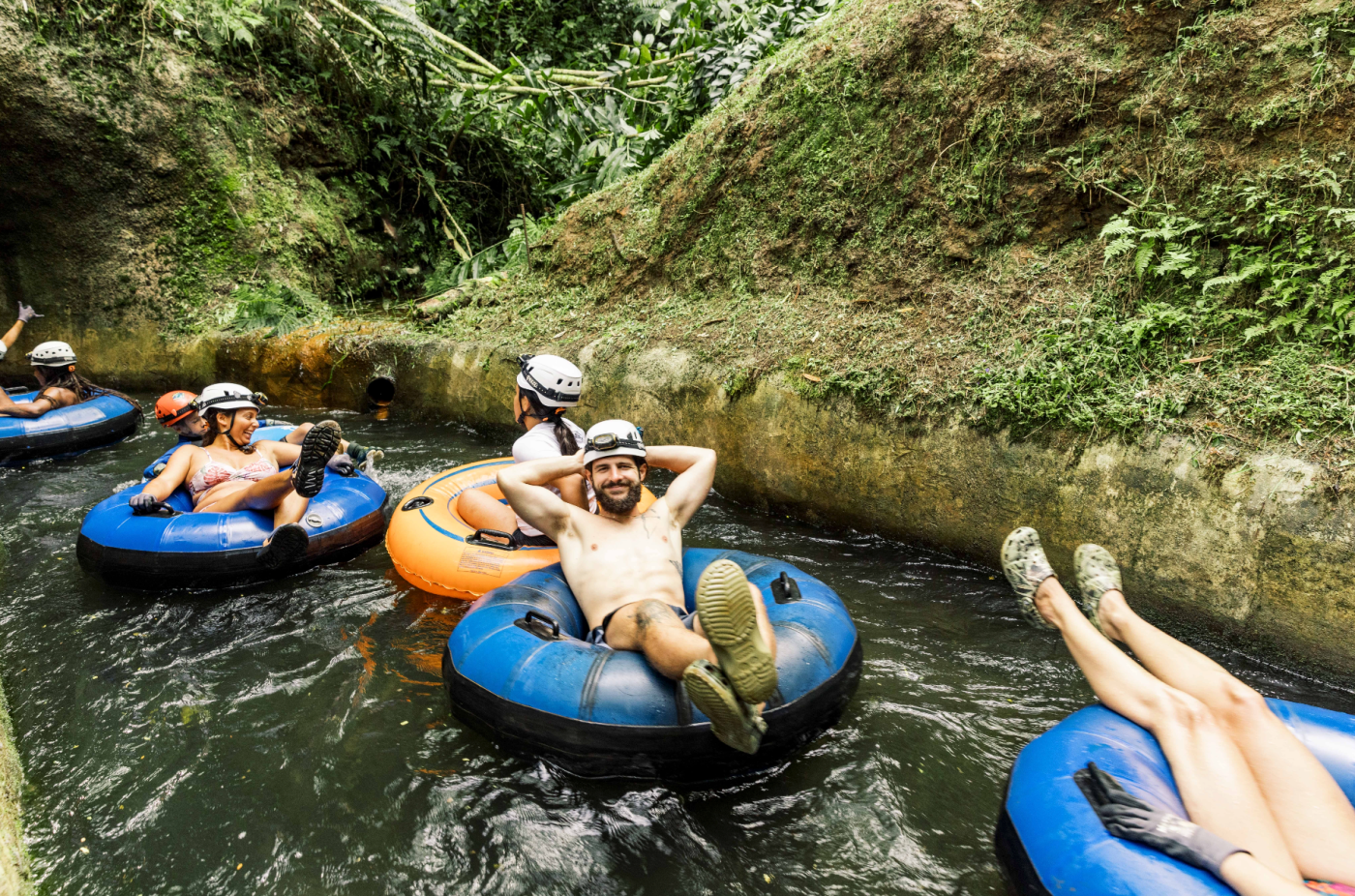People tubing in narrow stream with helmets and life jackets.