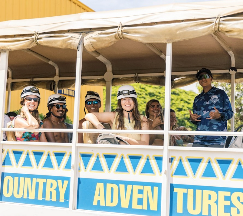 Group of people on a vehicle ride, wearing helmets, smiling and waving.