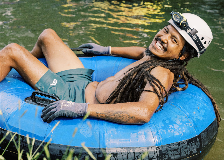 Person with dreadlocks in a helmet, smiling and reclining on a blue inner tube in water.
