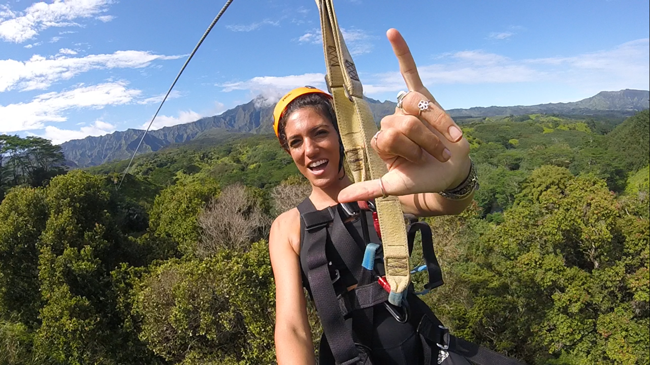 Person zip-lining over forested area, making a hand sign, with mountains in the background.