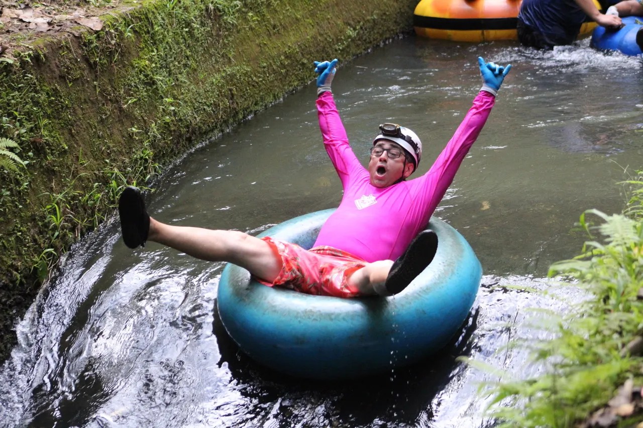 Person in pink shirt tubing down a narrow stream, arms raised, wearing a helmet and gloves.