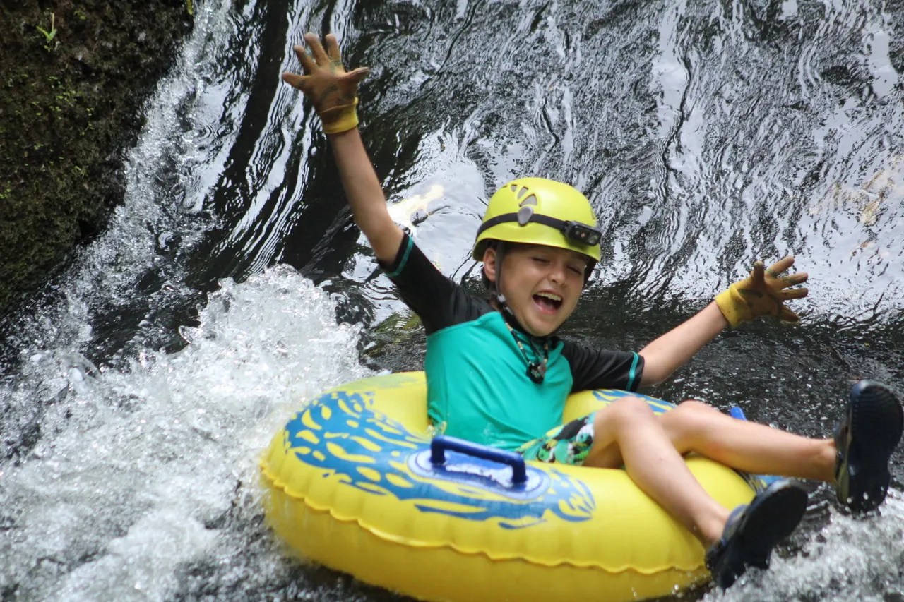 Child in helmet tubing down a waterfall, arms raised, smiling.