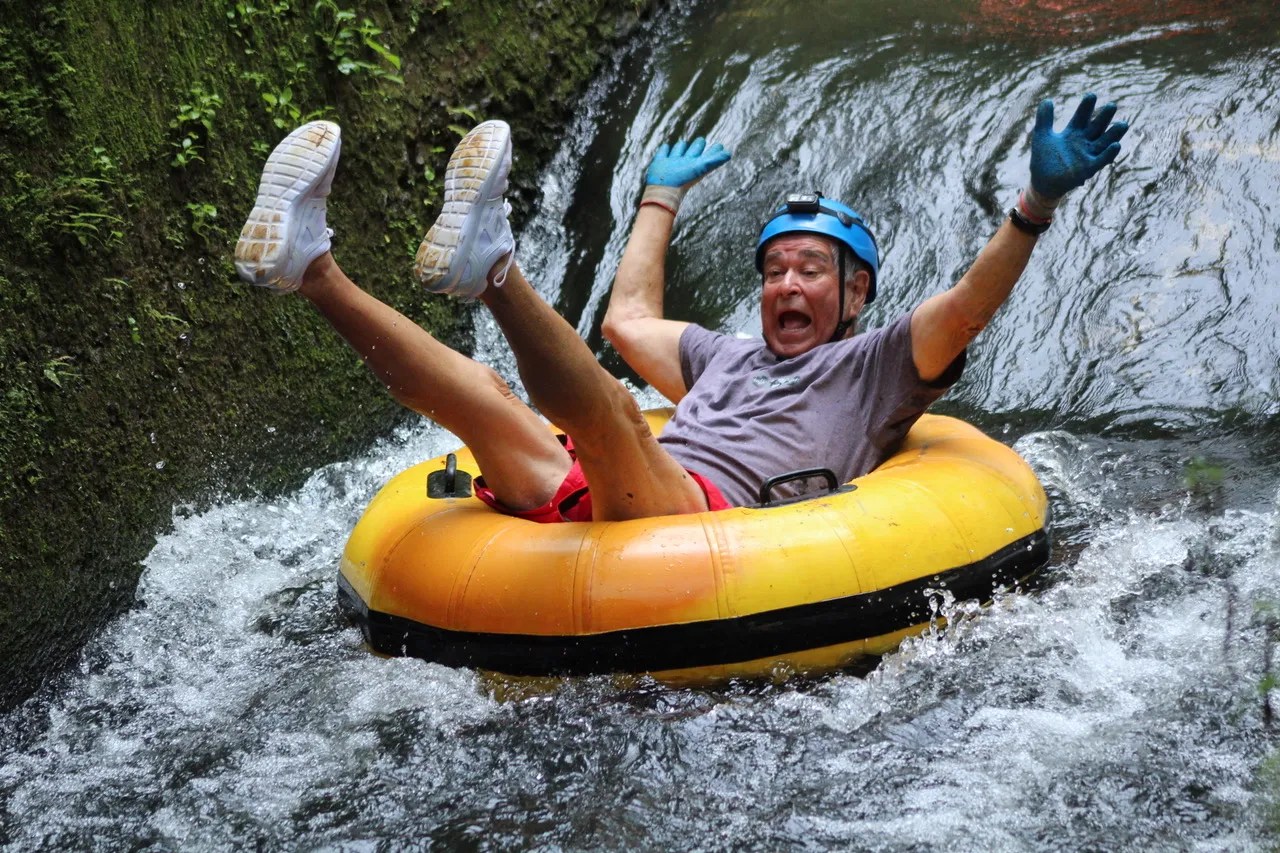 Person in helmet and gloves tubing down a water slide, arms raised in excitement.