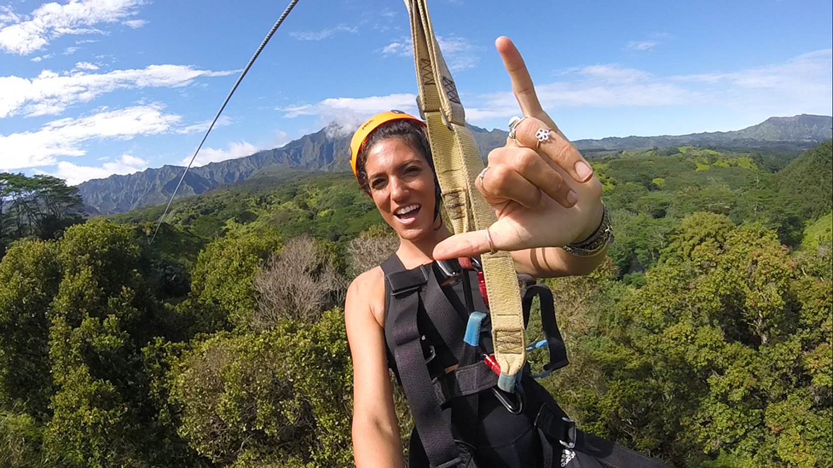 Person zip-lining over a forested area, gesturing with hand, mountain in the background.