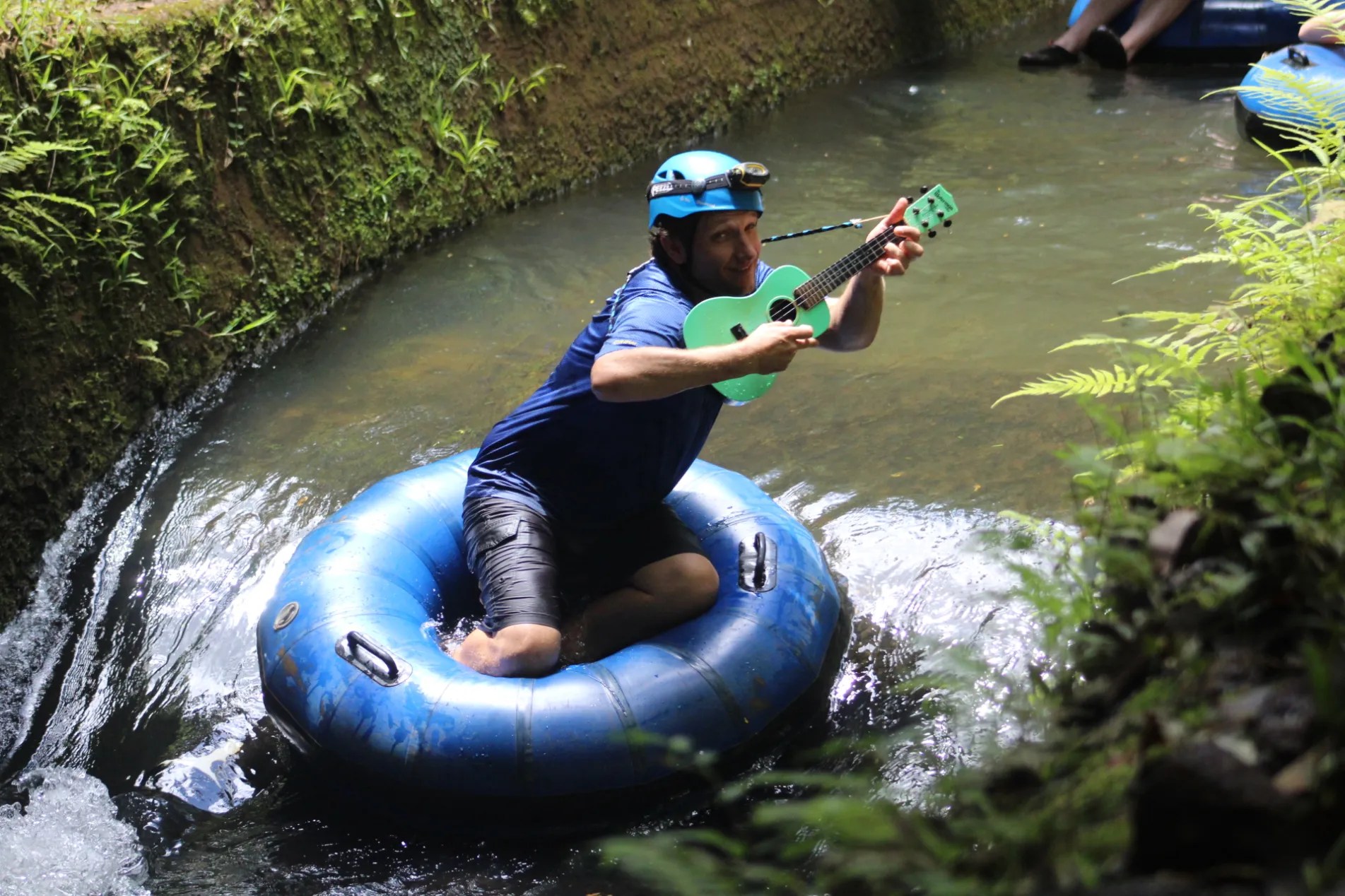 Man on a blue tube in a creek plays a green ukulele, wearing a blue helmet.