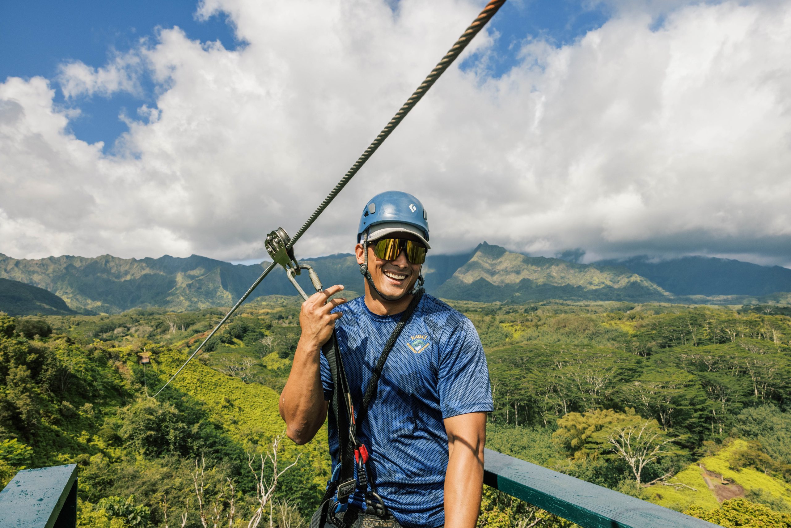 Man smiling while holding a zipline handle in lush mountain scenery.