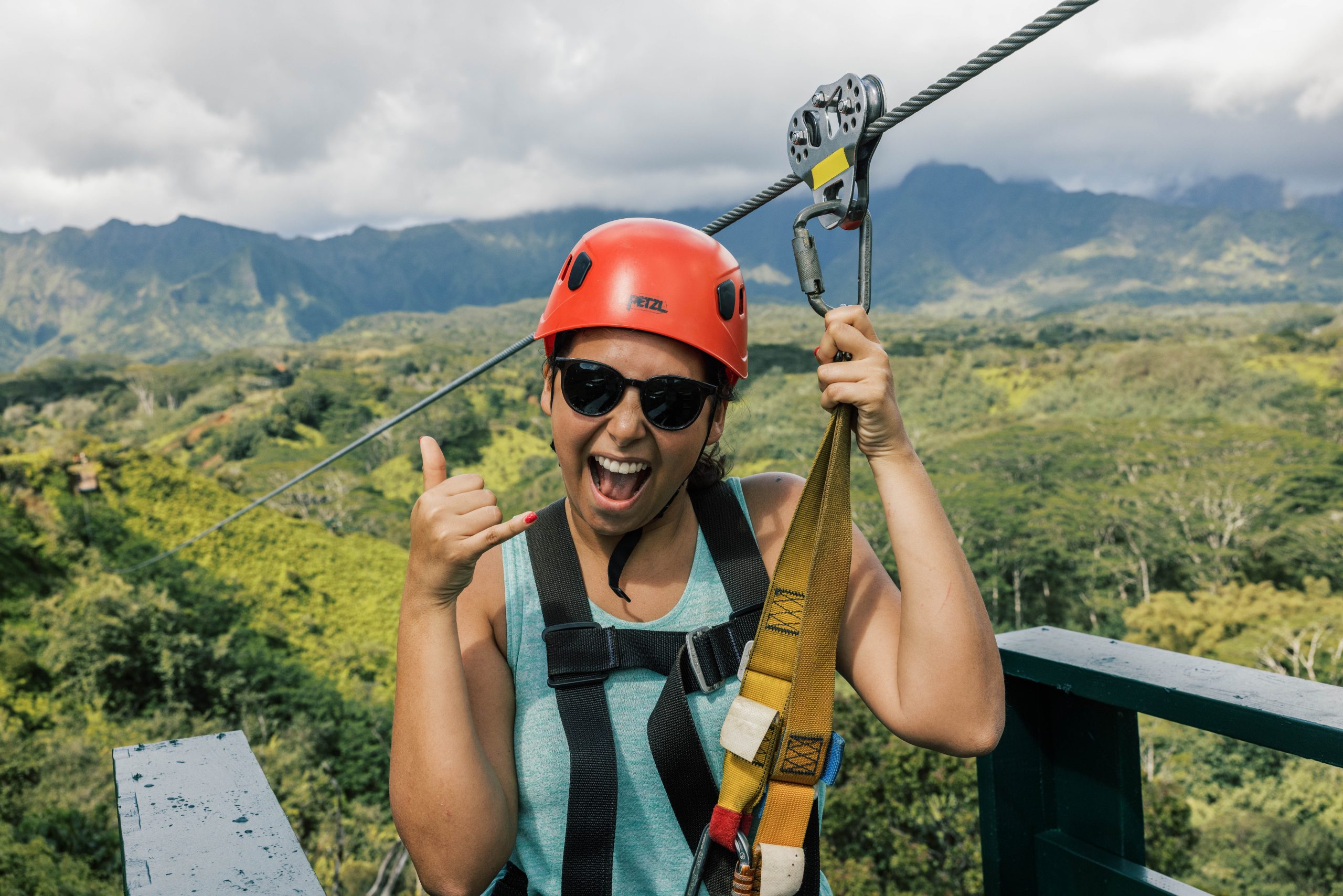 Person ziplining with a red helmet, sunglasses, and harness, giving a thumbs-up gesture.