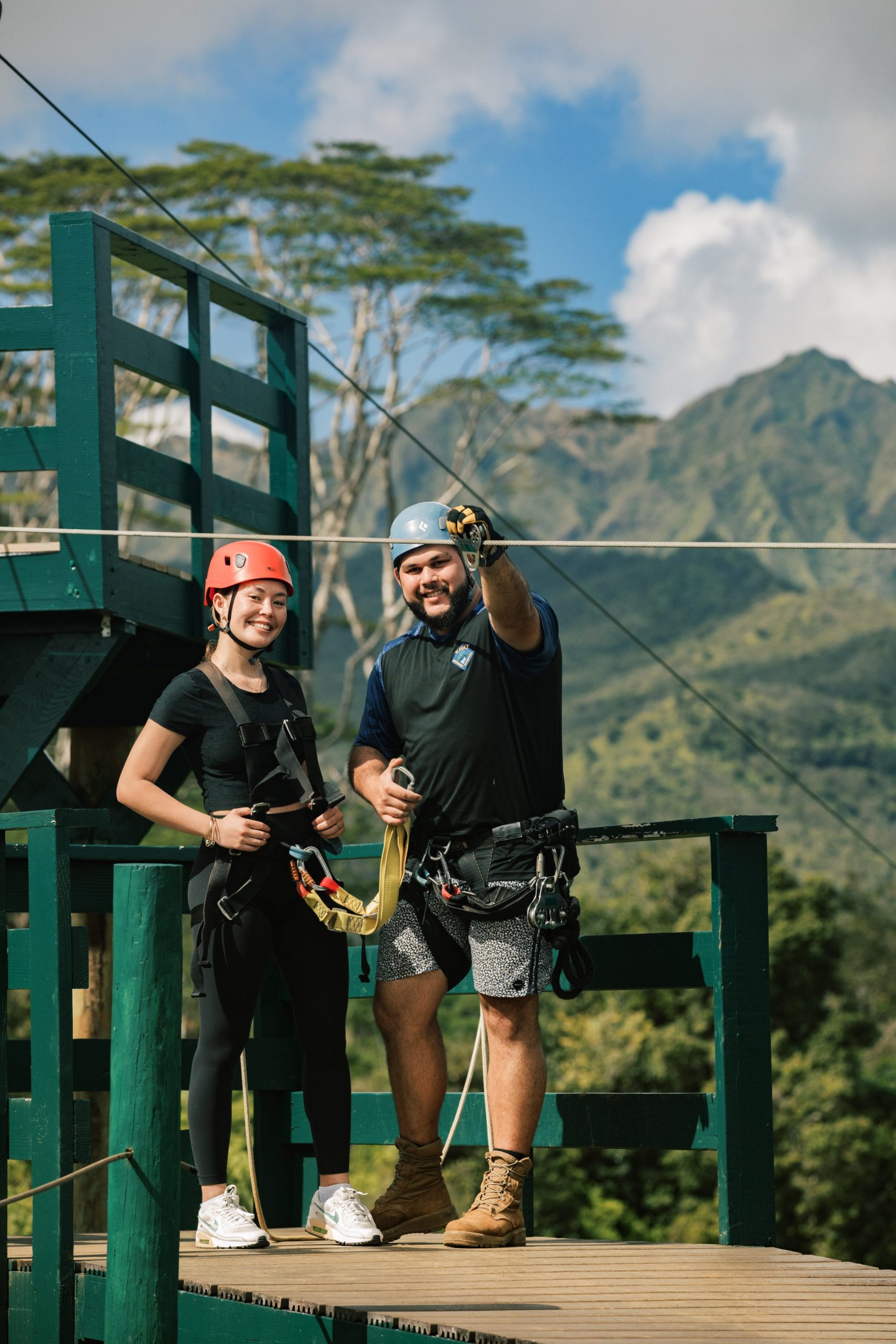 Two people in zip-lining gear on a wooden platform with mountainous backdrop.