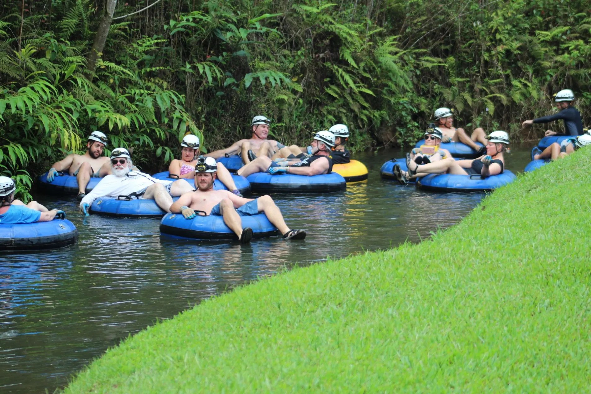Group tubing down a lush river, wearing helmets and life vests.