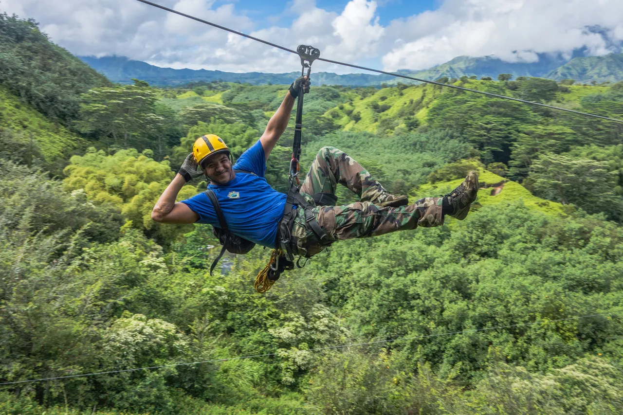 Person ziplining over lush green forest, wearing helmet and blue shirt.