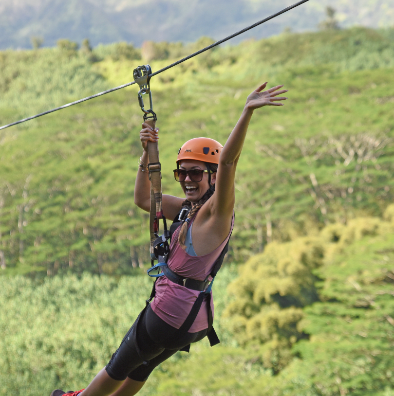Person ziplining over lush greenery, wearing a helmet and harness, waving.